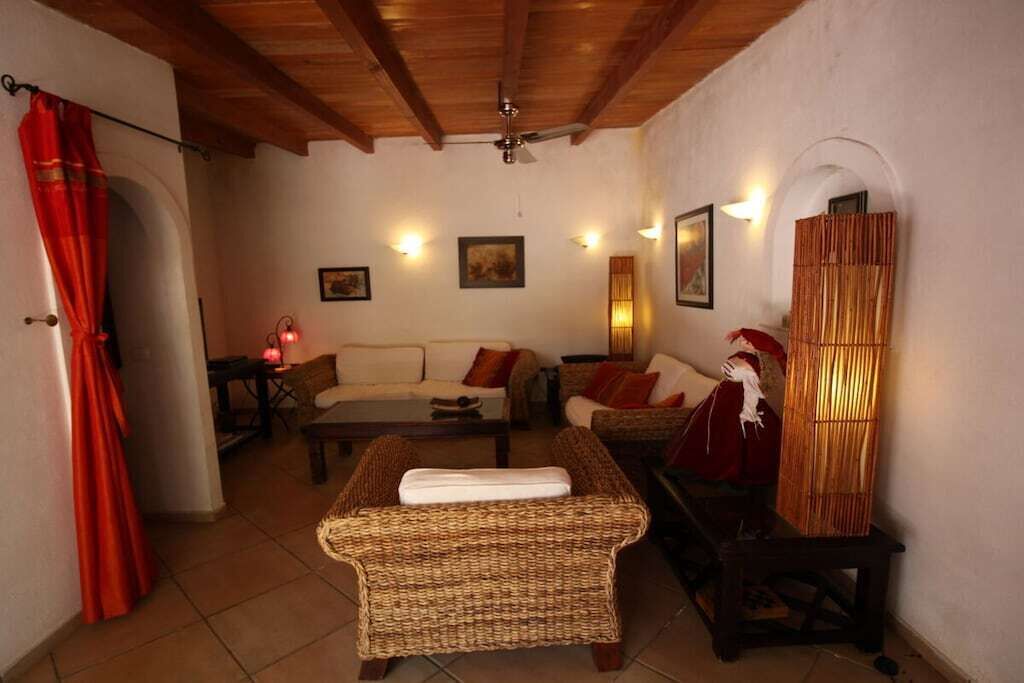 Un salon chaleureux avec un canapé, un fauteuil en osier et une table. On y trouve des lampes fixées au mur et un ventilateur de plafond.