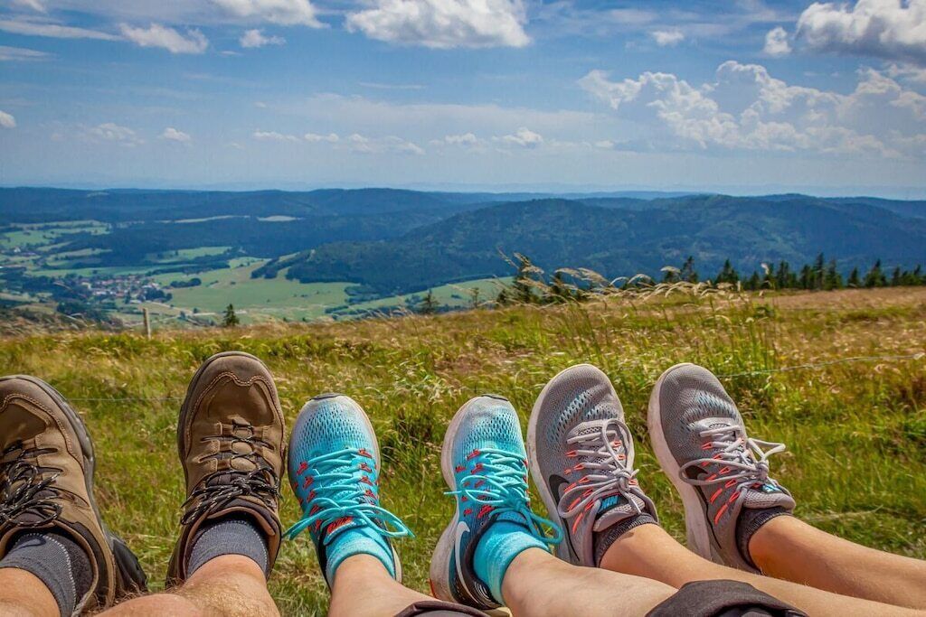 Piernas de una persona con botas de senderismo y zapatillas deportivas, descansando sobre la hierba con vistas a colinas distantes.