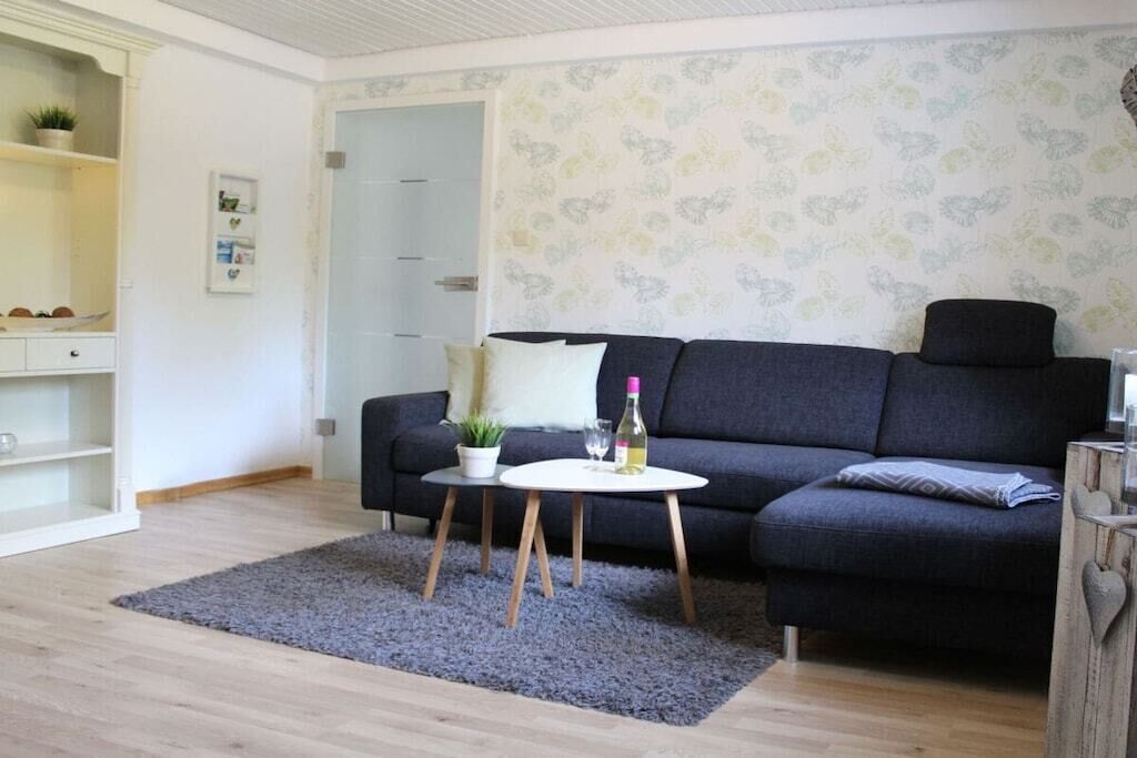 Een woonkamer met een donkergrijze bank, een klein rond tafeltje met een plant en een fles, en een grijze vloerkleed op een houten vloer.