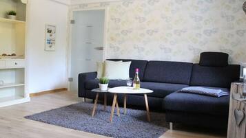 Een woonkamer met een donkergrijze bank, een klein rond tafeltje met een plant en een fles, en een grijze vloerkleed op een houten vloer.