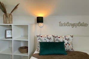 Een slaapkamer met een wit bed, een groen kussen, een wandlamp, een plankje met een mand, en een bordje met de tekst 'leblingsplatz'.