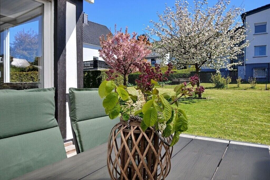 Une terrasse dotée de sièges confortables aux coussins verts, d’un centre de table en fibres tressées et d’un jardin planté d’arbres en fleurs.