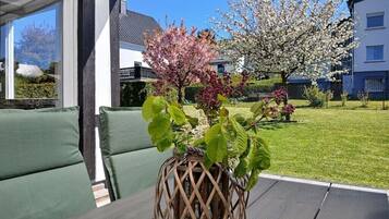 Une terrasse dotée de sièges confortables aux coussins verts, d’un centre de table en fibres tressées et d’un jardin planté d’arbres en fleurs.