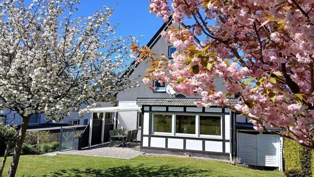 Uma casa moderna com fachada branca e grandes janelas, rodeada por um gramado bem cuidado e cerejeiras em flor.