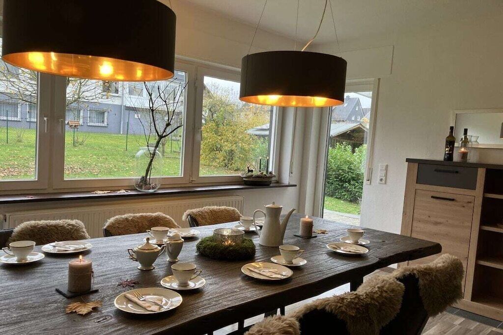 Un coin repas avec une table en bois dressée pour quatre personnes, entourée de chaises recouvertes de plaids en fourrure, une fenêtre donnant sur un jardin et des luminaires suspendus.