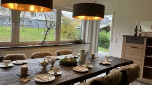 Uma sala de jantar com uma mesa de madeira posta para quatro pessoas, rodeada por cadeiras com mantas de pele, uma janela com vista para um jardim e iluminação pendente.
