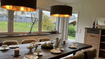 Un coin repas avec une table en bois dressée pour quatre personnes, entourée de chaises recouvertes de plaids en fourrure, une fenêtre donnant sur un jardin et des luminaires suspendus.