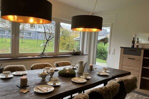 A dining area with a wooden table set for four, surrounded by chairs with fur throws, a window with a view of a garden, and pendant lighting. - Country House Moments in Olsberg (Olsberg)