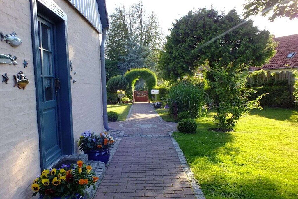 Un chemin mène à un bâtiment doté d’une porte bleue, lequel est entouré d’un jardin bien entretenu où poussent diverses plantes et fleurs.