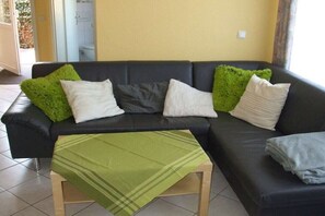 A black sofa with white and green pillows, a green throw blanket, and a wooden table in a room with tiled flooring and a yellow wall. - Kerstin Rehm holiday apartment (Cuxhaven)
