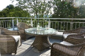 Een ronde glazen tafel omringd door rieten stoelen op een buitenbalkon met een metalen hekwerk.