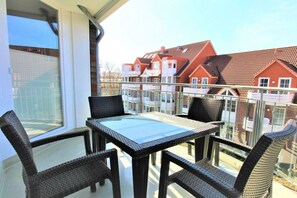Een balkon met een glazen tafel en rieten stoelen, met uitzicht op een rij rode huizen.