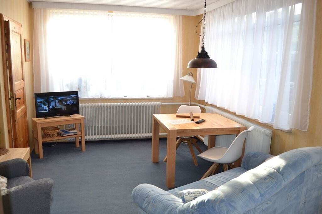 A room with a television on a wooden stand, a dining table with chairs, a radiator, and a window with curtains.