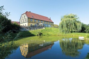A traditional half-timbered building with a red roof, a pond in the foreground, and a weeping willow tree to the right. - Holiday apartment with 1 bedroom (Gohrisch OT Papstdorf)
