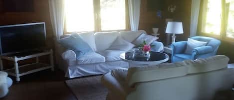 A living room with a white sofa set, a television on a stand, a coffee table with a lamp, and windows with curtains.