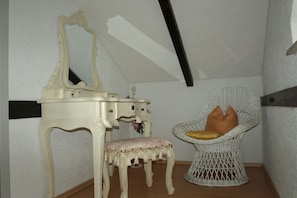 A room with a vanity with a mirror, a small stool, and a wicker chair with cushions.