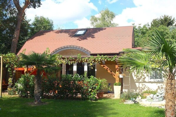 A house with a sloped roof, a covered porch, and a garden with various plants and a palm tree. - Mengelberg in the Leipzig New Lake District (Markkleeberg)