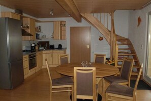 Une cuisine avec des meubles de rangement en bois, un réfrigérateur en acier inoxydable, une table à manger avec des chaises et un escalier en bois.