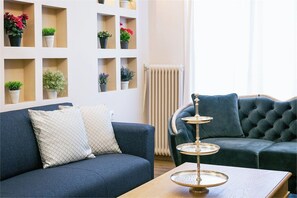 Ein Wohnzimmer mit einem dunkelblauen Sofa, einem weißen Kissen mit Muster, einem kleinen Tisch mit dekorativer Ablage und einer Wand mit mehreren Regalen, in denen Blumentöpfe stehen.