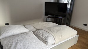 Un llit amb un llençol blanc i almoinos, un televisor de pantalla plana sobre un suport, i una finestra amb cortines.