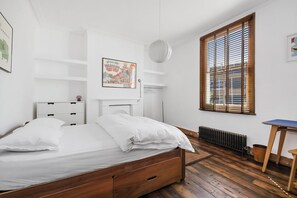 Ein Schlafzimmer mit einem hölzernen Bett, weißen Wänden, einem Heizkörper, einem Fenster mit Jalousien und einem kleinen Tisch.