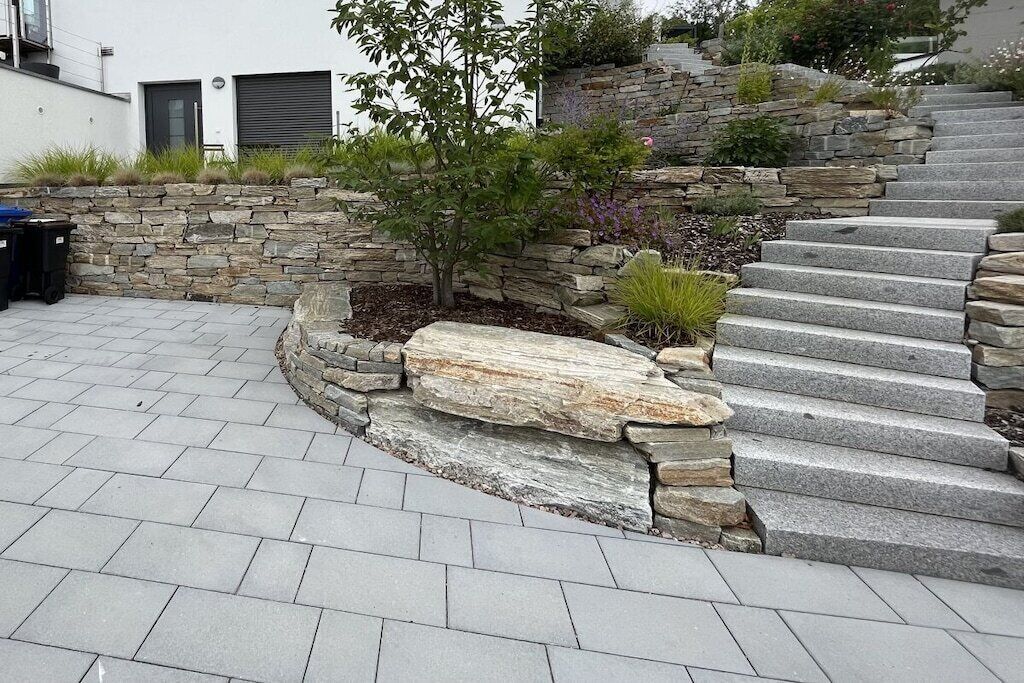 Een stenen trap leidt naar een gebouw met een modern design, omringd door een aangelegde tuin met verschillende planten.