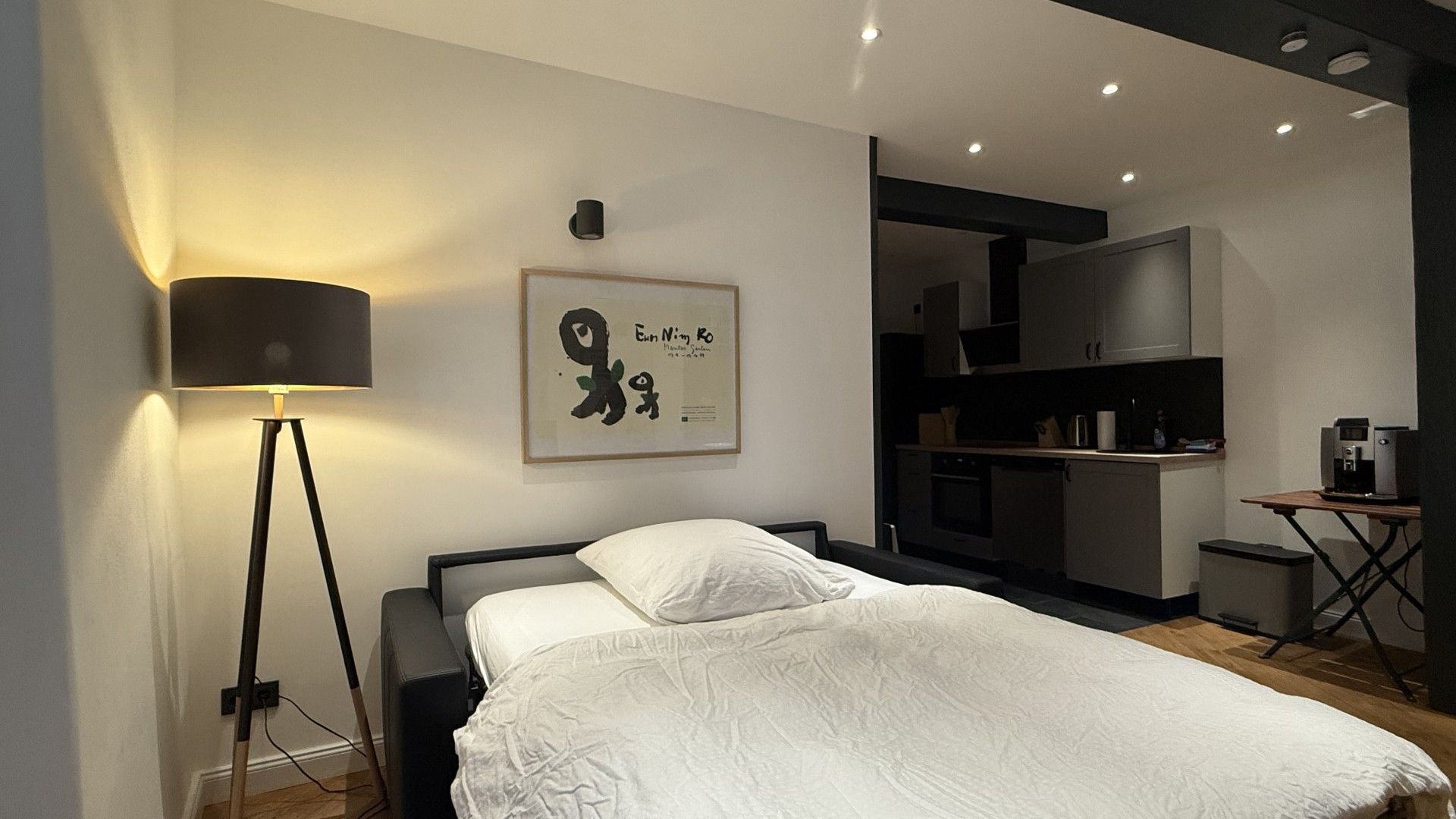 Ein modernes Schlafzimmer mit einem Bett, einer Nachttischlampe, einem gerahmten Kunstwerk und einer Küchenzeile im Hintergrund.