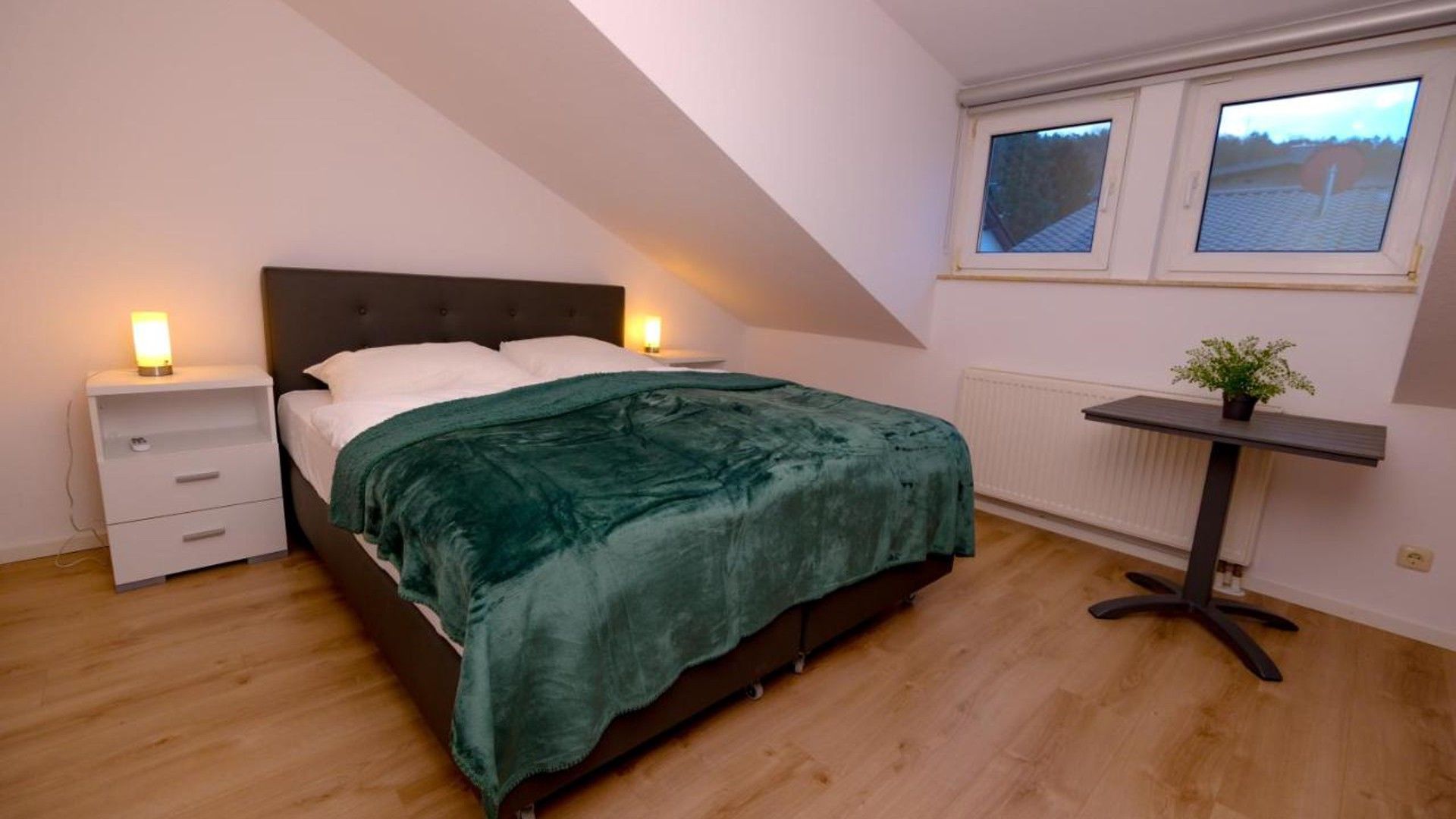 Ein Schlafzimmer mit einem Bett, Nachttischen, einem Heizkörper, einem Fenster und einem kleinen Tisch mit einer Pflanze.