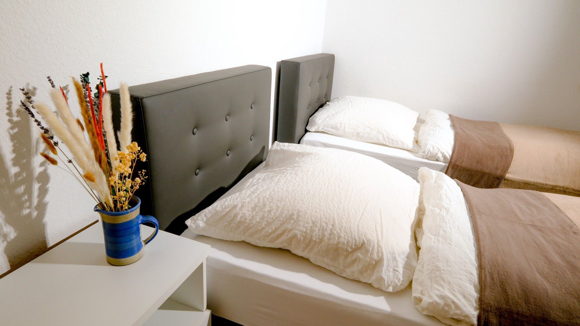 A bedroom with a grey upholstered headboard, white bedding, and a vase with dried flowers on a bedside table.