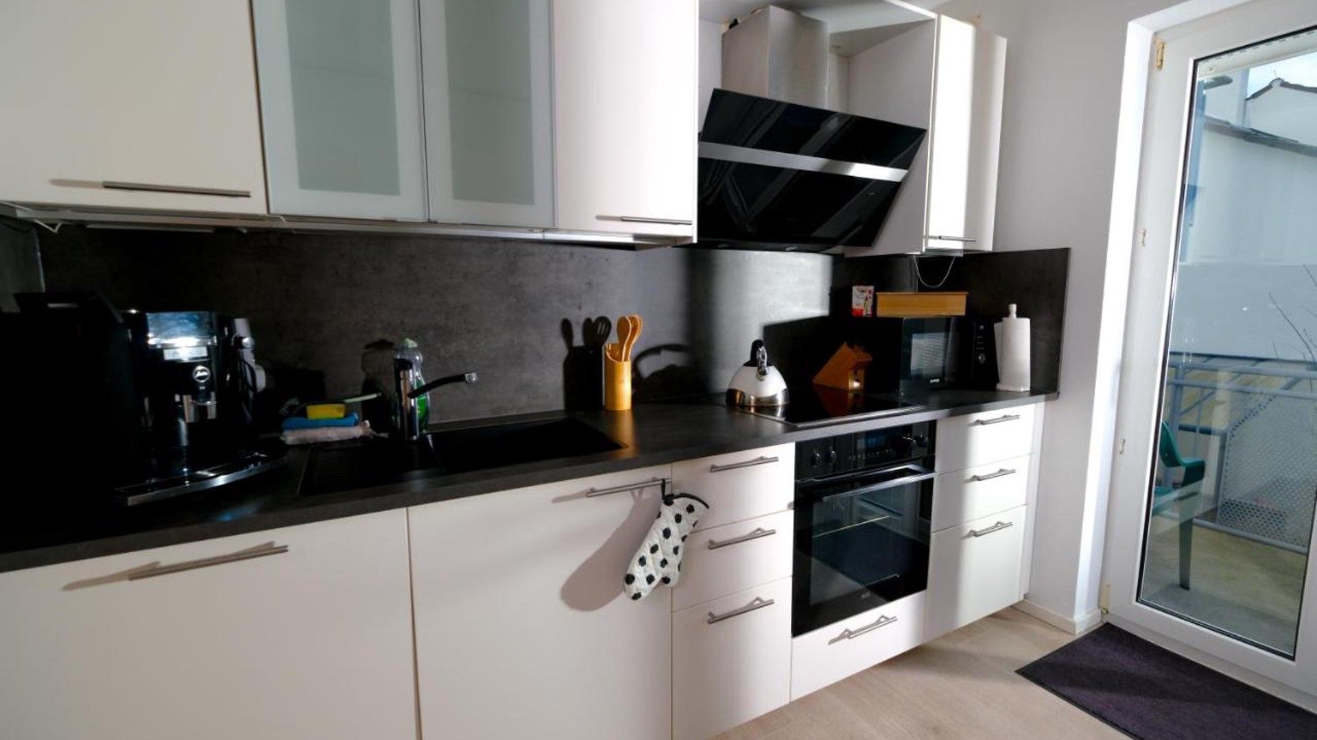 A modern kitchen with white cabinets, a black countertop, and stainless steel appliances including a coffee maker, kettle, and microwave.