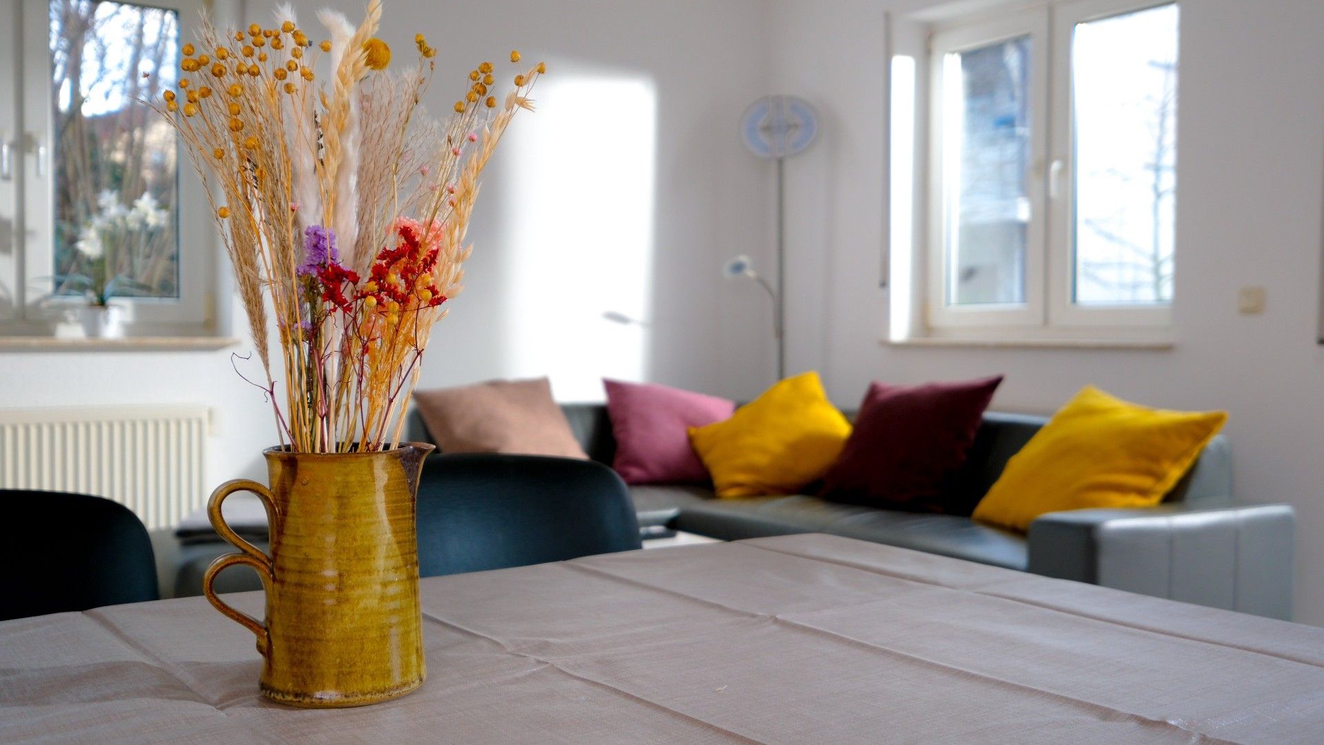 A living room with a table, a vase with dried flowers, and a sofa with colorful cushions.