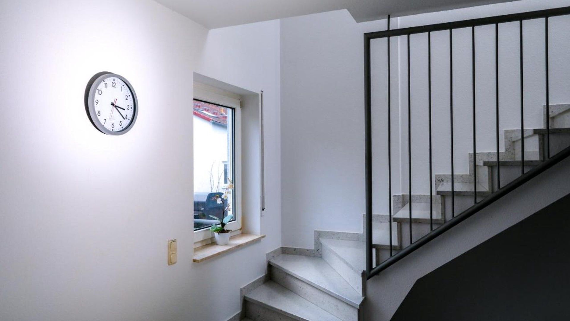 A staircase with a black railing leading to a landing, a wall-mounted clock, and a window with a view of the outside.
