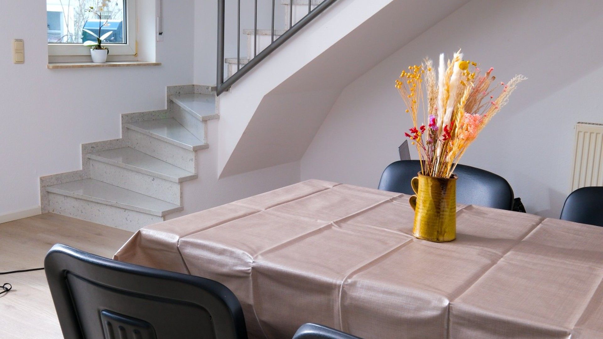 A dining area with a table covered in beige tablecloth, a vase with dried flowers, and a staircase in the background.