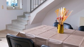 Ein Essbereich mit einem Tisch, der mit einer beigen Tischdecke bedeckt ist, einer Vase mit getrockneten Blumen und einer Treppe im Hintergrund.