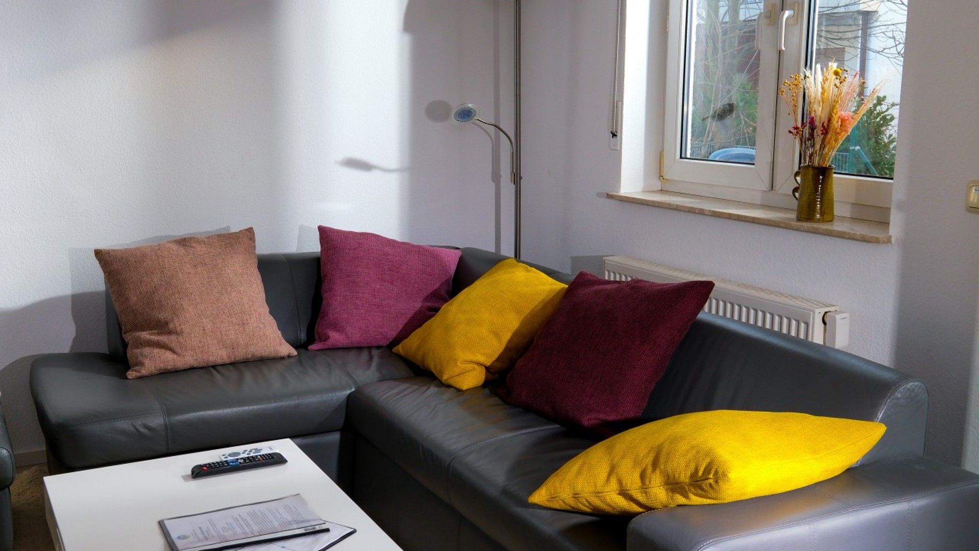 Ein modernes Wohnzimmer mit einem grauen Sofa, einem weißen Couchtisch und einem Fenster mit Blick auf Grünflächen.