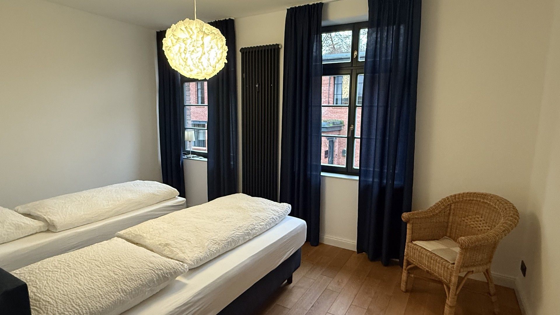 A room with two beds, a wicker chair, a hanging light fixture, and a window with curtains.