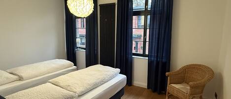 Una habitación con dos camas, una silla de mimbre, una lámpara colgante y un ventanal con cortinas.