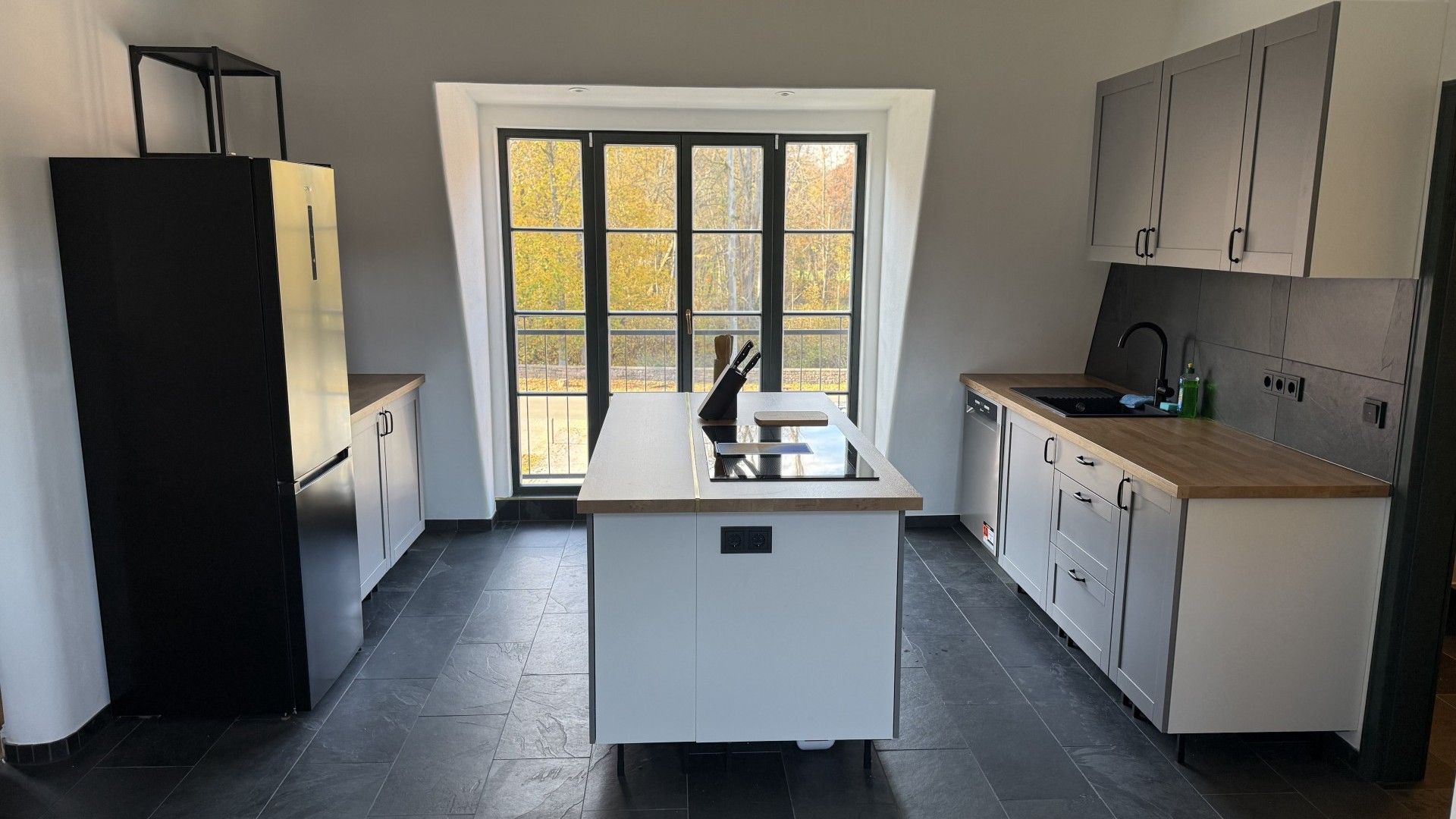 A modern kitchen with a central island, a refrigerator, and a window with a view of trees.