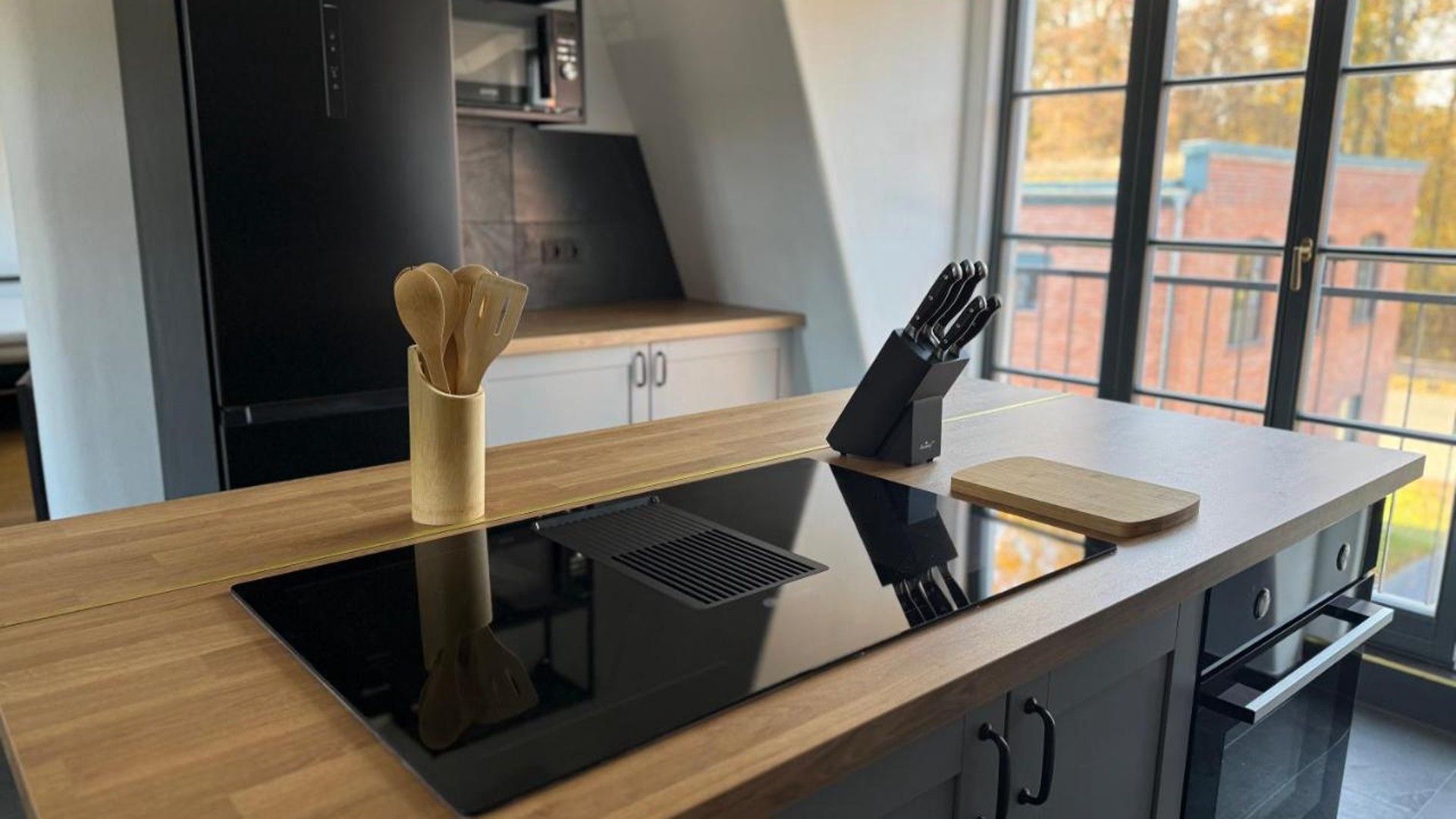 Une cuisine moderne dotée d’une plaque de cuisson à induction noire, de meubles en bois, d’un bloc à couteaux et d’une fenêtre donnant sur l’extérieur.