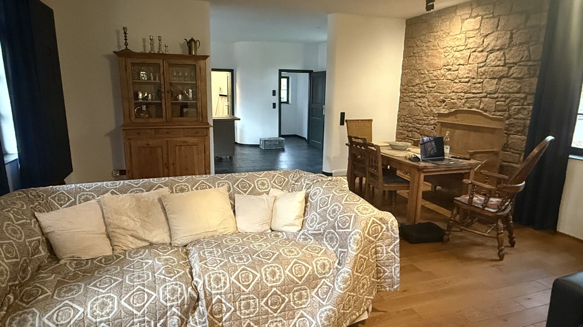 Una habitación con un sofá estampado, un armario de madera, una mesa de comedor con sillas y una pared de piedra.
