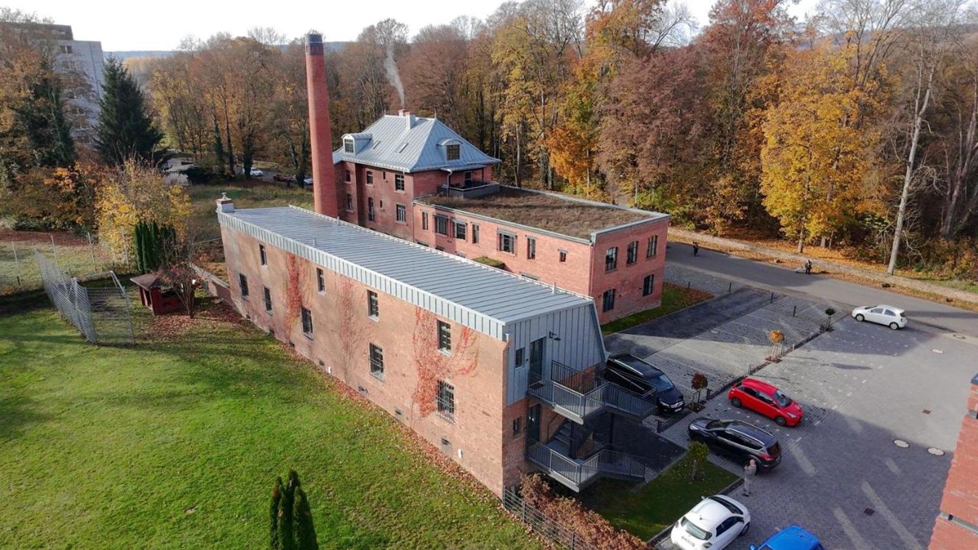 A large industrial building with multiple floors, a prominent chimney, and a parking area with several cars.
