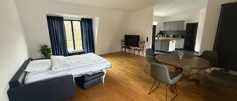 A modern hotel room with a bed, a dining table, a TV, and a kitchenette.