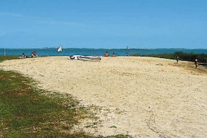 Una playa arenosa con un velero, gente y un muelle distante.