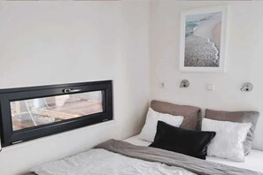 Un dormitorio con cama, almohadas, una ventana y una lámpara de pared.