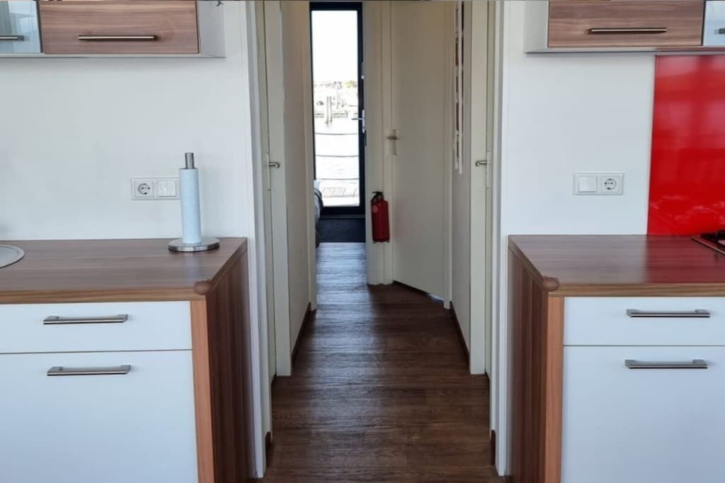 Un couloir étroit doté d’un plancher en bois, de meubles blancs aux poignées argentées et d’un mur d’accent rouge.