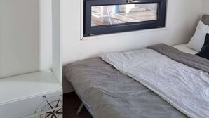 Een eenpersoonsbed met een grijs matras en witte beddengoed, een klein raam met een zwarte lijst, en een witte nachtkastje met een kompas dessin.