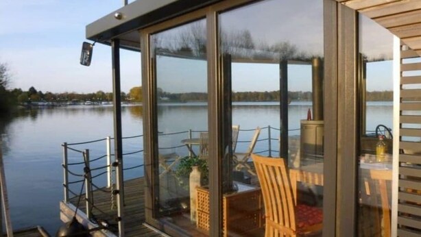 Een glazen terras met uitzicht op een meer, houten meubels en een tafel met een fles.