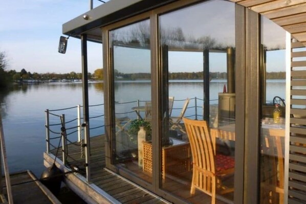 A glass-enclosed deck with a view of a lake, wooden furniture, and a table with a bottle. - Houseboat in Anjum with swimming pool (Anjum)