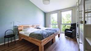 Ein Schlafzimmer mit einem hölzernen Bett, einem Stuhl und einem Schreibtisch mit Sitzgelegenheit. Zwei Fenster sorgen für Tageslicht und bieten Blick ins Grüne.