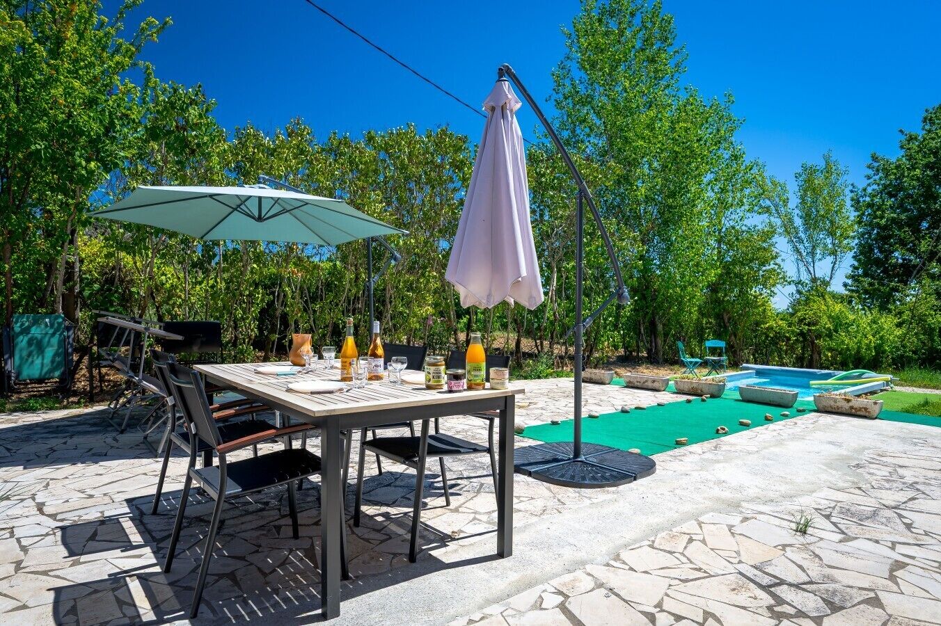 Ein Essbereich im Freien mit Tisch, Stühlen und Sonnenschirmen, umgeben von einem Schwimmbecken und Grünflächen.